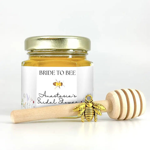 Small jar of honey with personalized Bride to Bee label, gold bee charm, and wooden honey dipper on white background