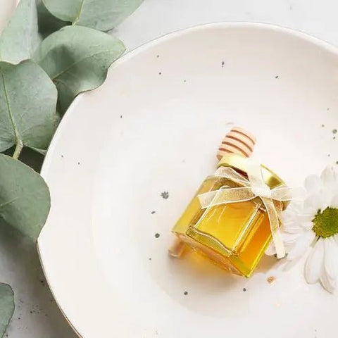 Small Jar of honey with gold lid and honey dipper laying on white plate on white surface with greenery