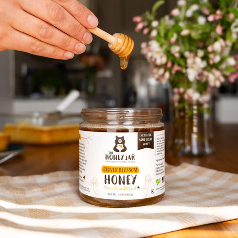 Hand holding a honey dipper above a Jar of honey sitting on a table with a gingham towel and flowers