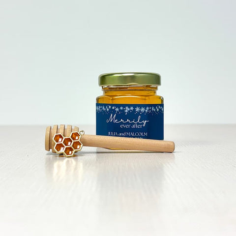 Jar of honey with a blue label, honeycomb charm, and wooden honey dipper on a white background