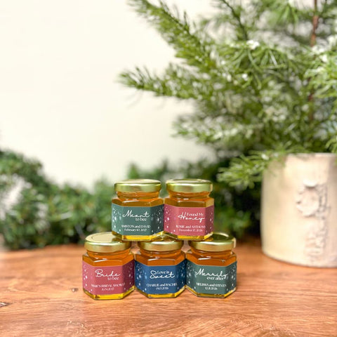 Five jars of honey with labels on a wooden surface with a blurred green plant background