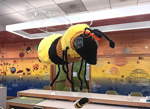 Plush bee with sunglasses hanging from ceiling over a table against a colorful wall.