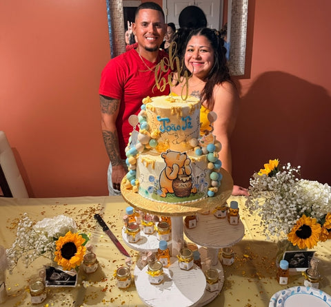 Couple standing behind a baby shower cake with Winnie the Pooh design, surrounded by flowers and small bottles of honey.