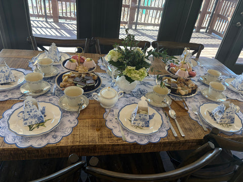 Dining table set for a tea party with teacups, cakes, and floral arrangements.
