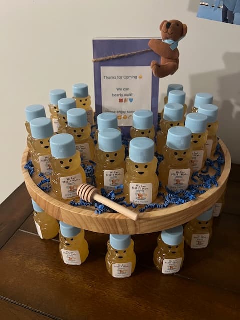 Display of small bear-shaped honey bottles with blue lids on a wooden stand, accompanied by a teddy bear and a sign.