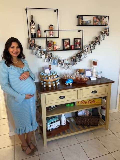 Pregnant woman in a blue dress standing next to a decorative shelf with various items.