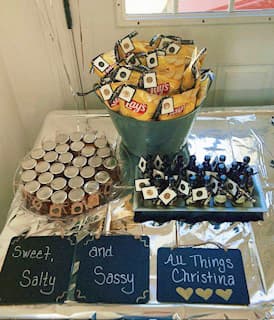 Table with snacks and decorative items including a bouquet of snacks, honey jars, and signs labeled 'Sweet and Salty' and 'All Things Christina'.