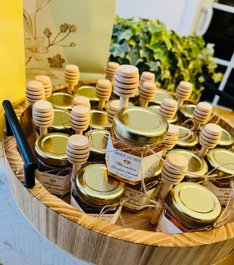 Jars of honey with wooden dipper sticks and personalized labels on a wooden tray.