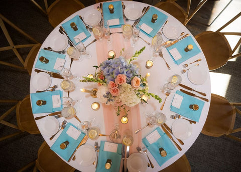 Round table set for a formal event with blue place mats, gold cutlery, small honey jars with gold lids, and a floral centerpiece.