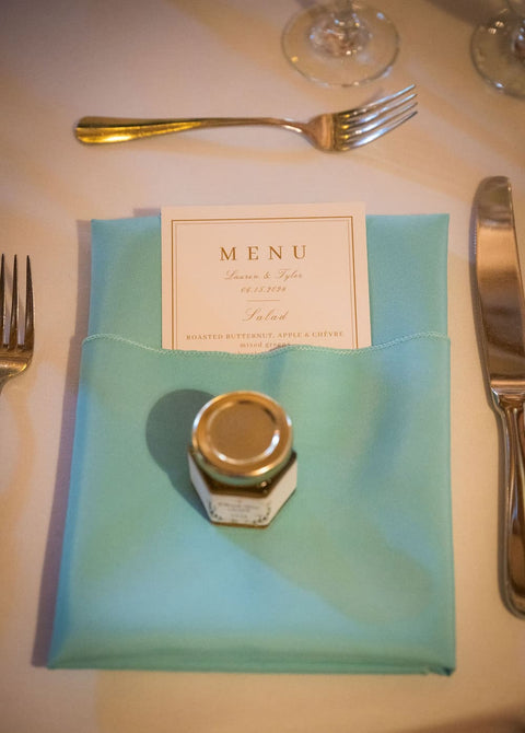 Teal napkin with a menu card and small jar of honey on a table setting with cutlery.