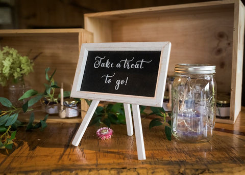 Small chalkboard sign on a wooden stand with 'Take a treat to go!' text, next to a mason jar on a wooden surface.