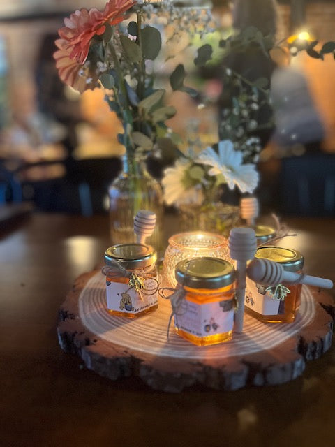 Three jars of honey on a wooden board with flowers and a candle in the background.
