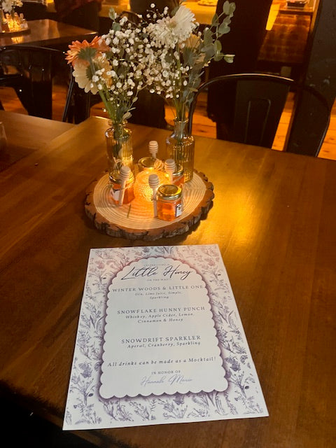 Decorative table setting with flowers, small bottles of honey, and an invitation card on a wooden surface.