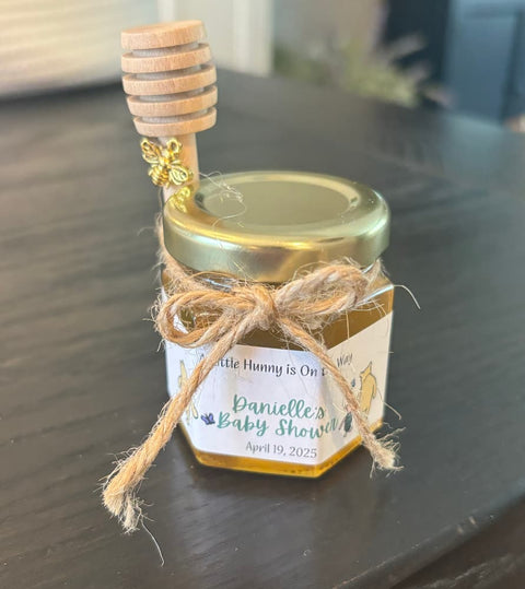 Small jar of honey with a wooden dipper on a dark surface, tied with twine and labeled for a baby shower.