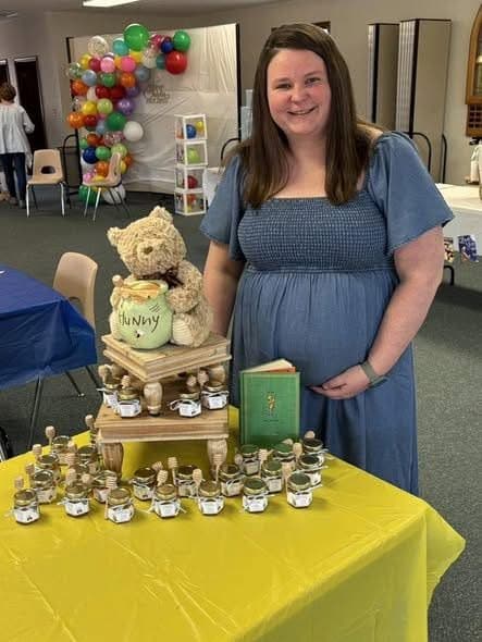 Pregnant woman standing behind a table with baby shower decorations, including teddy bears and jars.