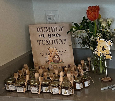 Honey jars and honey dippers with a sign and floral arrangement on a countertop.