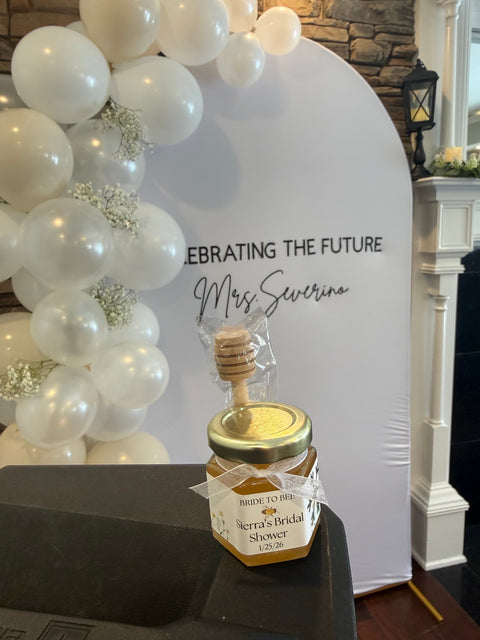 Jar of honey with a dipper on a table in front of a decorative backdrop with balloons.