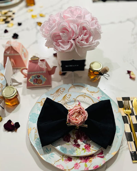 Decorative table setting with floral centerpiece, black bow, small honey jars, and floral napkin on a white tablecloth.