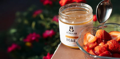 Jar of honey with a spoon dripping honey into a bowl of strawberries, flowers in the background