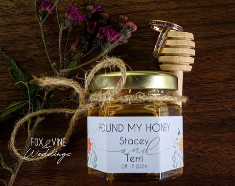 Jar of honey with a wooden dipper and floral arrangement on a wooden surface, featuring a personalized label.