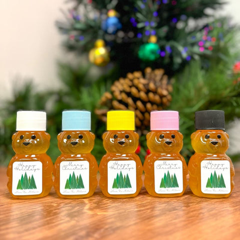 Five bear-shaped honey jars with colorful lids on a wooden surface with a Christmas tree in the background.