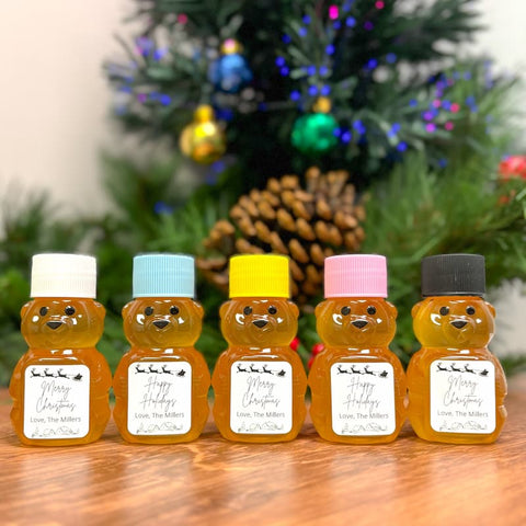 Five bear-shaped honey bottles with colorful lids in front of a decorated Christmas tree.
