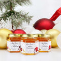Three jars of honey with Christmas-themed labels in front of festive decorations.