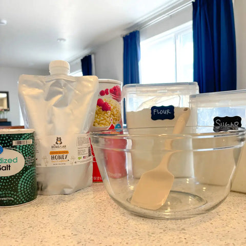 Kitchen counter with baking ingredients and utensils including a glass bowl, measuring cups, Mylar pouch of honey, and containers.
