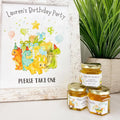 Decorative sign for a birthday party with dinosaur illustrations, accompanied by honey jars with personalized labels.