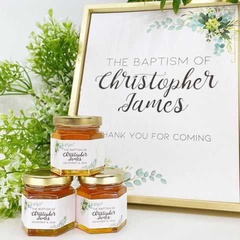 Three jars of honey with personalized labels in front of a framed sign for a baptism celebration.