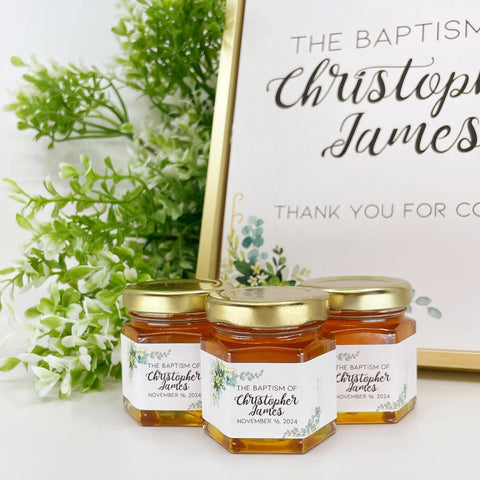 Three honey jars with personalized labels next to a decorative sign with greenery.