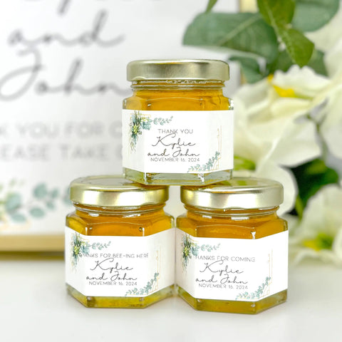 Three small jars of honey with personalized labels on a white surface with greenery in the background.