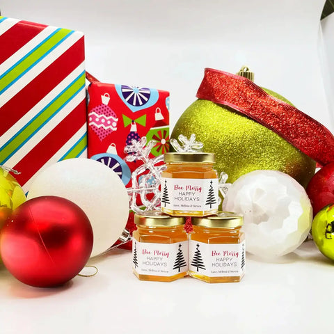 Three jars of 'Bee Merry' honey with Christmas ornaments and presents in the background.