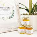 Jars of honey with baby shower favors on a white surface with a plant and framed text in the background.