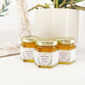 Three small jars of honey with gold lids and personalized labels on a white surface