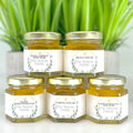 Five jars of honey with personalized labels on a white surface with green plants in the background.