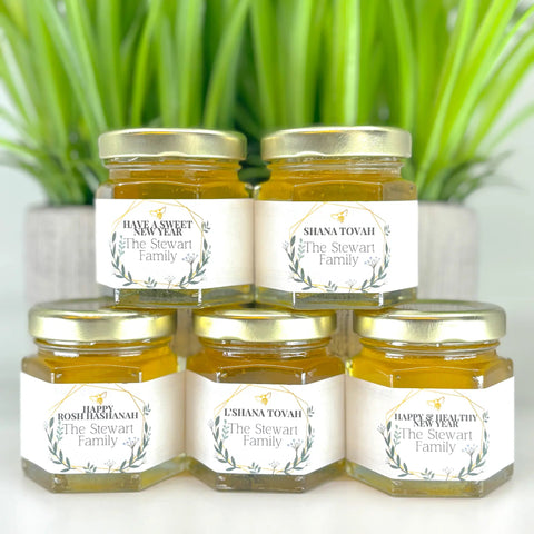 Five jars of honey with personalized labels on a white surface with green plants in the background.