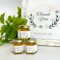 Three jars of honey with 'Thank You' labels on a white surface with greenery and a 'Thank You' sign in the background.