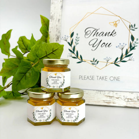 Three jars of honey with 'Thank You' labels on a white surface with greenery and a 'Thank You' sign in the background.