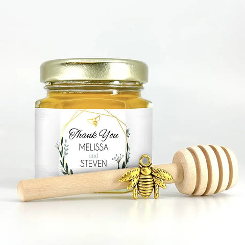 Jar of honey with a gold lid, gold bee charm, and wooden dipper on a white background
