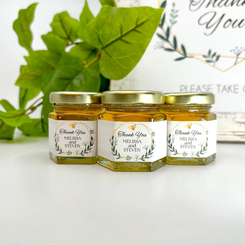 Three jars of honey with personalized labels on a white surface with greenery in the background.