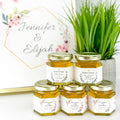 Jars of honey with personalized labels on a white surface with a plant in the background.