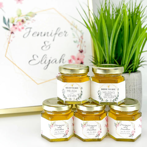 Jars of honey with personalized labels on a white surface with a plant in the background.