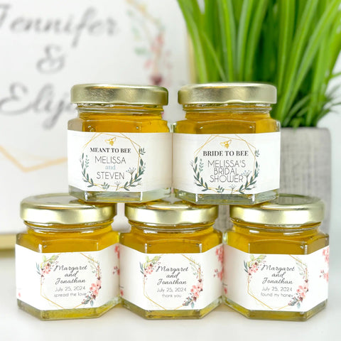 Jars of honey with personalized labels on a white surface with a plant in the background.
