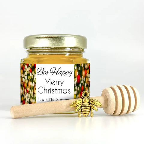 Jar of honey with a decorative label, a wooden honey dipper, and a gold bee charm on a white background.