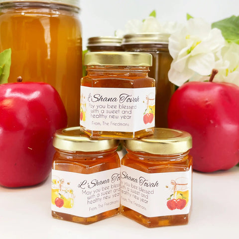 Jars of honey with Shana Tovah labels surrounded by apples and flowers on a white background