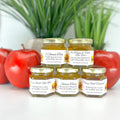 Five jars of honey with labels in front of red apples and a plant in the background