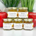 Five jars of honey with personalized labels on a white surface with red apples and green plants in the background.