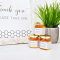 Honey jars with personalized labels on a white surface with a plant in the background.