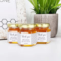 Three honey jars with personalized labels on a white surface with a plant in the background.